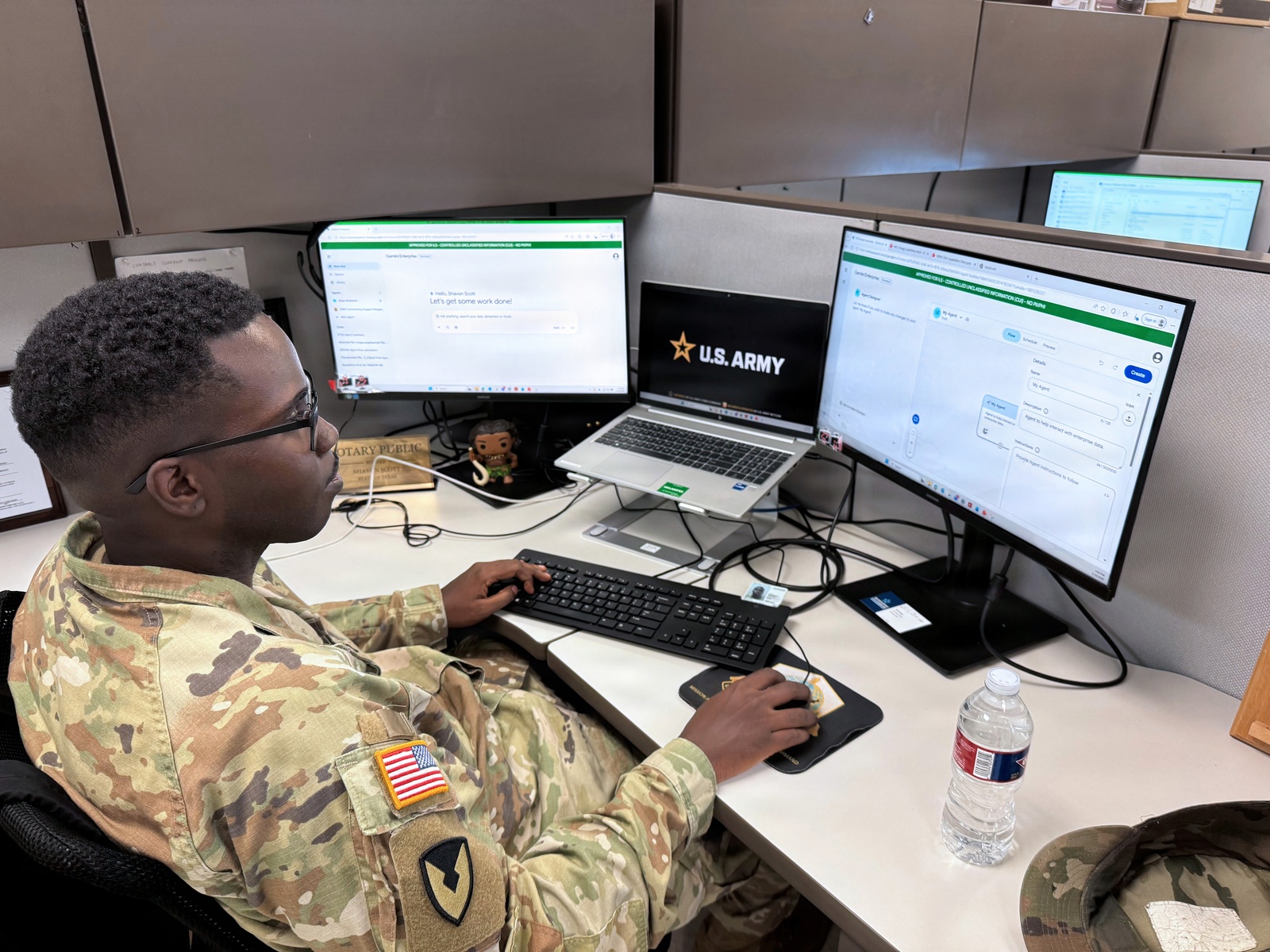 An Army contracting officer working at a desk with a laptop and two monitors