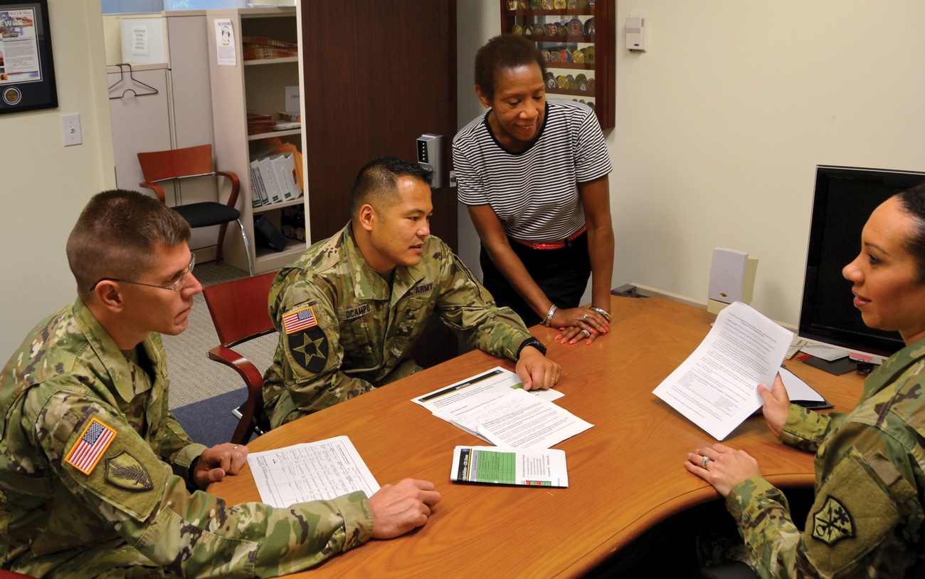 Four Army acquisition staff members reviewing printed documents around an office table