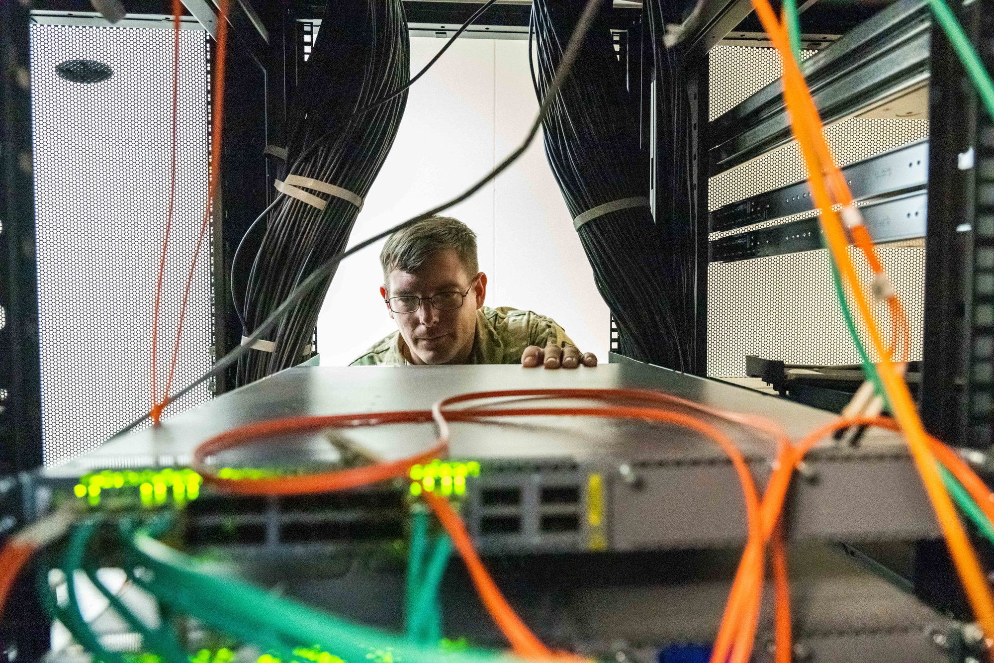 A uniformed technician inspects equipment between server racks and bundles of network cables.