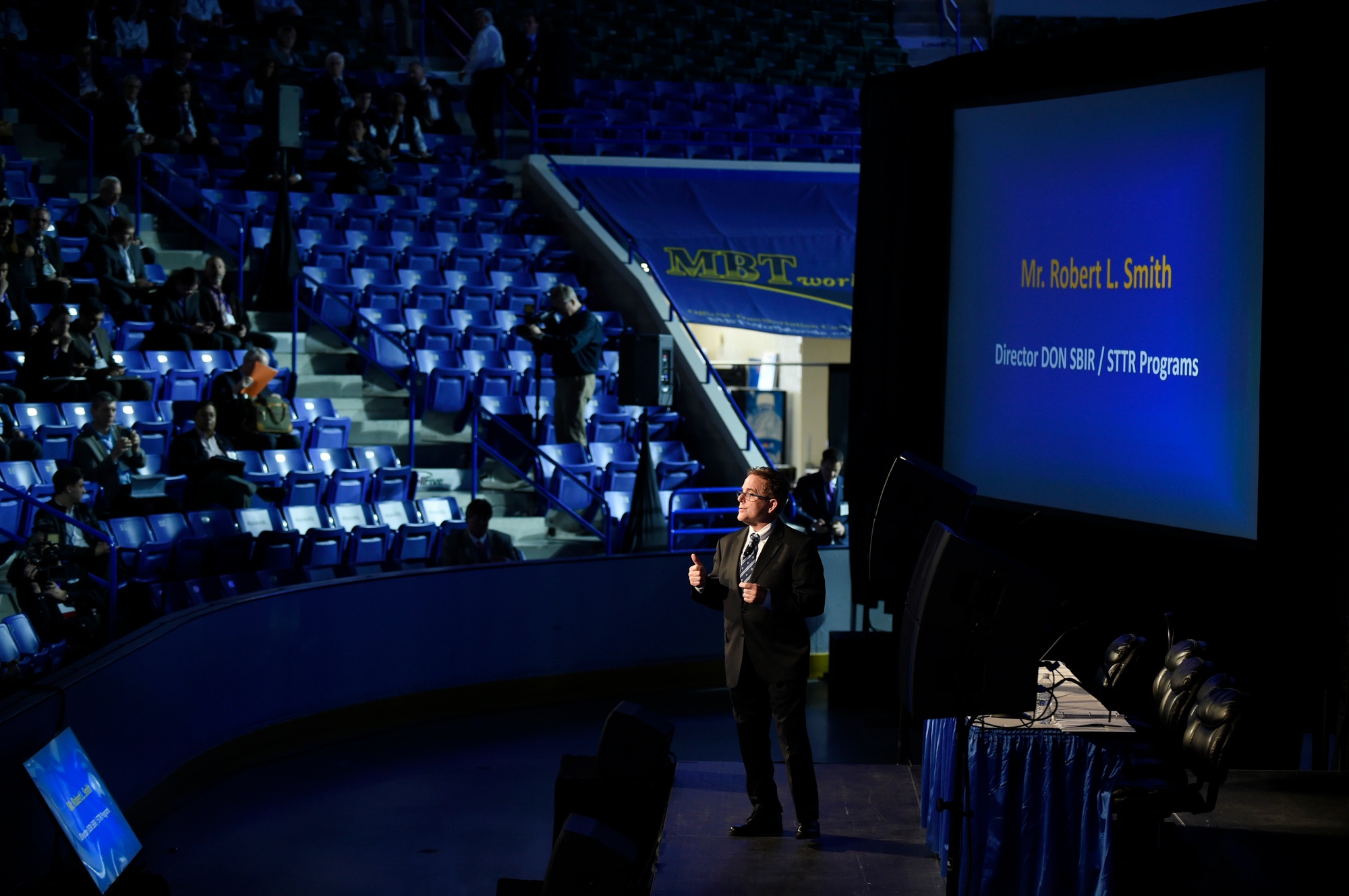 Speaker addresses attendees beside a screen identifying the Navy SBIR and STTR programs at the 2019 transition forum